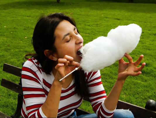 Woman eating an all-natural, dye-free cotton candy cone, sitting on a bench in a park.