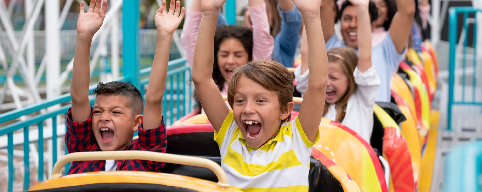 Children and adults on a roller coaster with arms raised, at an amusement park.